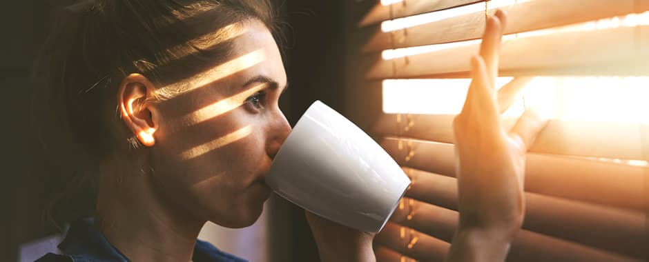 Woman looking out of faux wood blinds at the sun