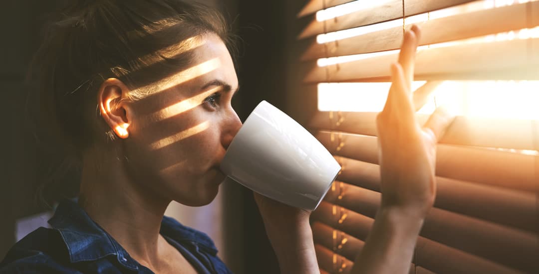 Woman looking out of faux wood blinds at the sun