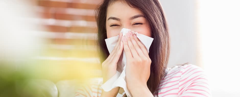 Woman sneezing into tissue in front of window blinds